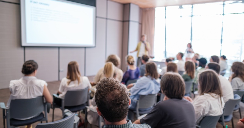 Attendees seated in a conference room listening to a presenter deliver a professional session, with a presentation slide projected on a screen.