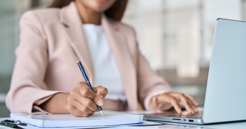 Professional woman in a business suit taking notes in a notebook while working on a laptop, symbolizing post-conference reflection and integration.