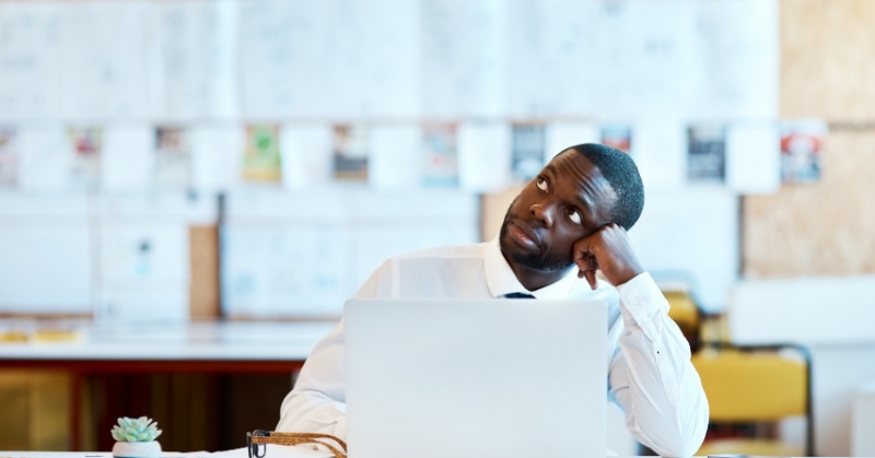 A man in business attire sits at a desk with a laptop, resting his head on his hand and gazing upward thoughtfully, suggesting contemplation or uncertainty in a modern office setting.