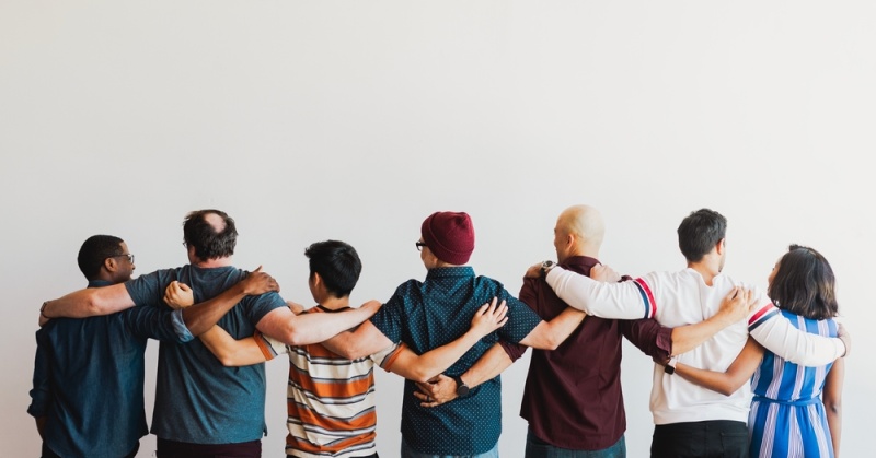 A diverse group of people with their arms around each other looking away from the camera.