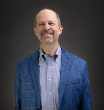 Joe Woodard, host of Scaling New Heights and CEO of Woodard, wearing a blue checkered blazer and smiling against a dark background.