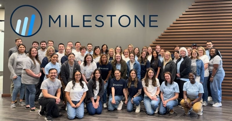 Group photo of the Milestone Business Solutions team posing indoors in front of a gray wall with the Milestone logo, featuring over 40 smiling team members in casual attire.