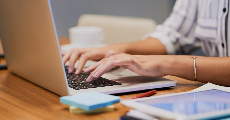 Close-up of a person typing on a laptop at a wooden desk, with a tablet, sticky notes, and a pen nearby.