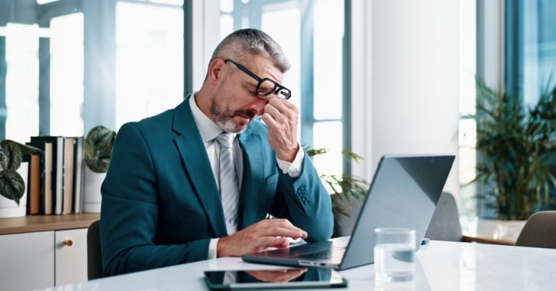Frustrated businessman in a teal suit rubbing his eyes while working on a laptop in a modern office, symbolizing tax season stress and 1099 deadline pressure.