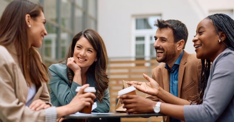 Four professionals sitting at an outdoor table, smiling and talking over coffee, representing connection and casual business relationships.