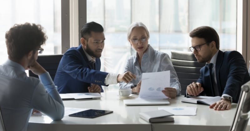 Four professionals in business attire sit around a conference table in a modern office, reviewing charts and documents during a focused team discussion.