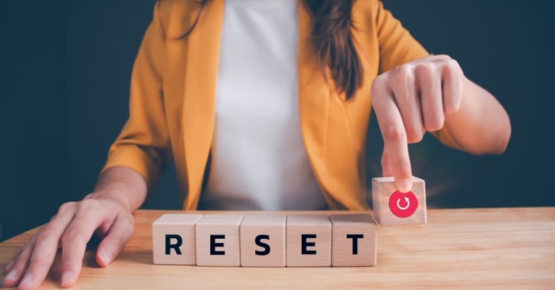 A person wearing a yellow blazer is seated at a table, pressing a wooden block with a red power icon. Four other wooden blocks spell the word "RESET," symbolizing a fresh start or new beginning.