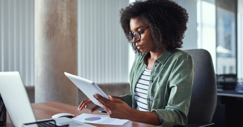 A woman wearing glasses sits at a desk in a modern office, reviewing a tablet while a laptop and printed chart rest in front of her.
