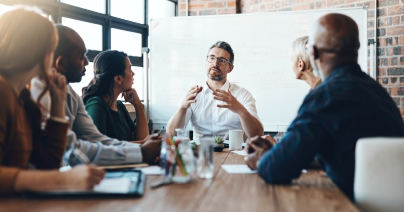 A business team in a meeting, gathered around a conference table, listening to a man speaking and gesturing in front of a whiteboard with strategy notes.