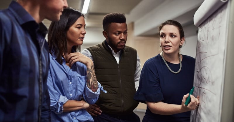 diverse group of professionals gathered around a flip chart, attentively listening as a woman leads a discussion and points to a diagram, illustrating leadership and collaboration in a business setting.