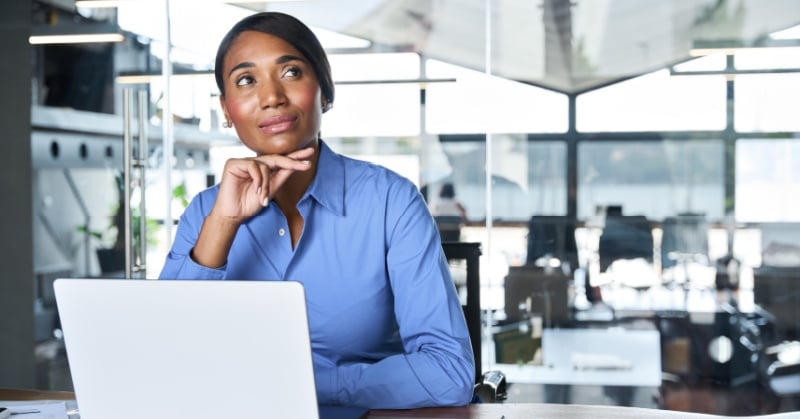 A professional woman in a blue button-down shirt sits at a desk in a modern office, gazing thoughtfully to the side with her hand resting on her chin. A laptop is open in front of her, suggesting contemplation or strategic planning.