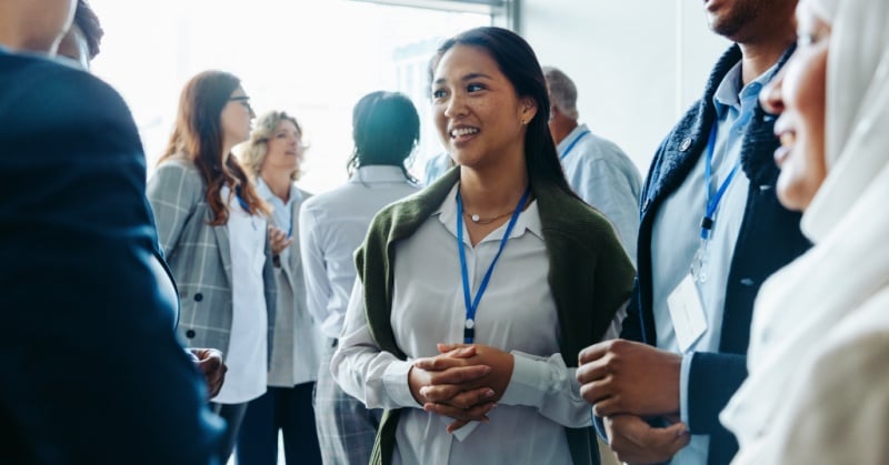 A diverse group of professionals networking at a business conference, with one woman in focus smiling and engaging in conversation, wearing a name badge and professional attire.