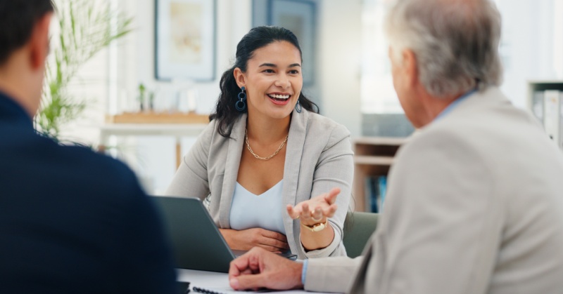A smiling professional woman leads a conversation at a meeting table with two colleagues in a modern office setting.