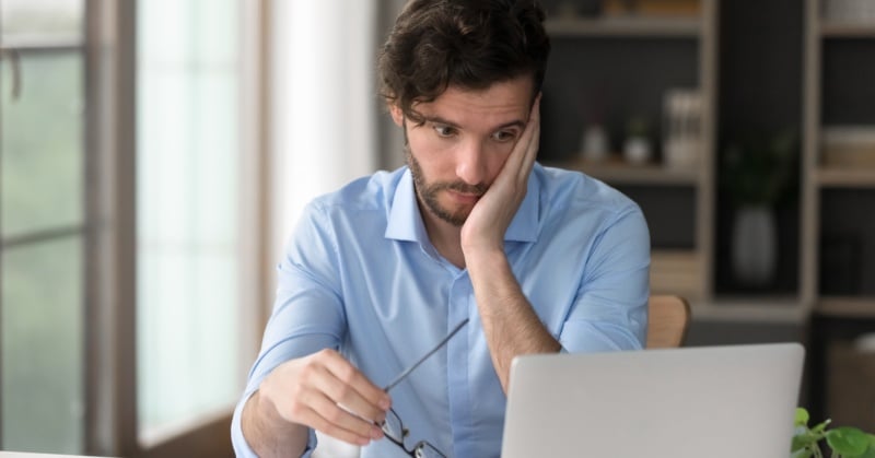 Stressed accountant looking at laptop screen while holding eyeglasses, overwhelmed by digital clutter in a modern office setting.