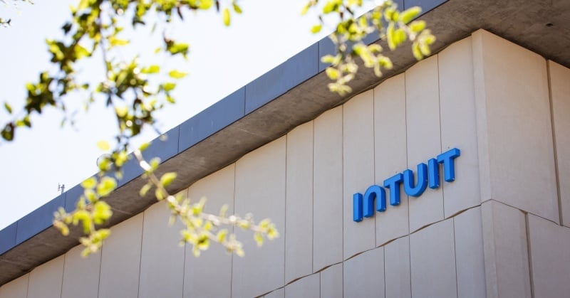 The exterior of an Intuit office building with the company’s blue logo mounted on a beige concrete wall, partially framed by blurred green tree branches.