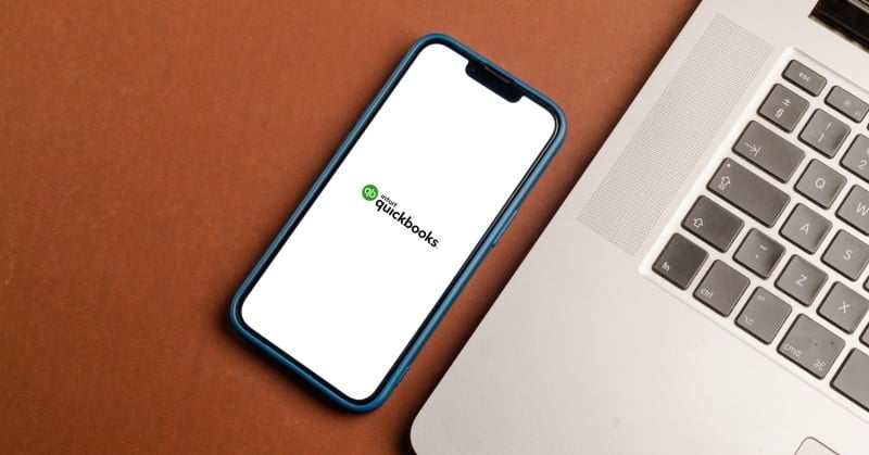 Smartphone displaying QuickBooks Online logo next to a laptop on a brown desk.