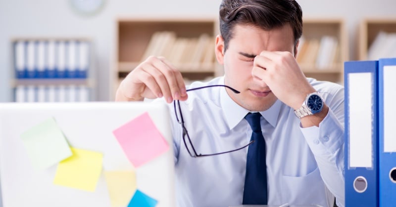 A stressed office worker sits at a desk, holding his glasses and rubbing his eyes in frustration, surrounded by folders, sticky notes, and a laptop, suggesting fatigue or overwhelm from work inefficiency.