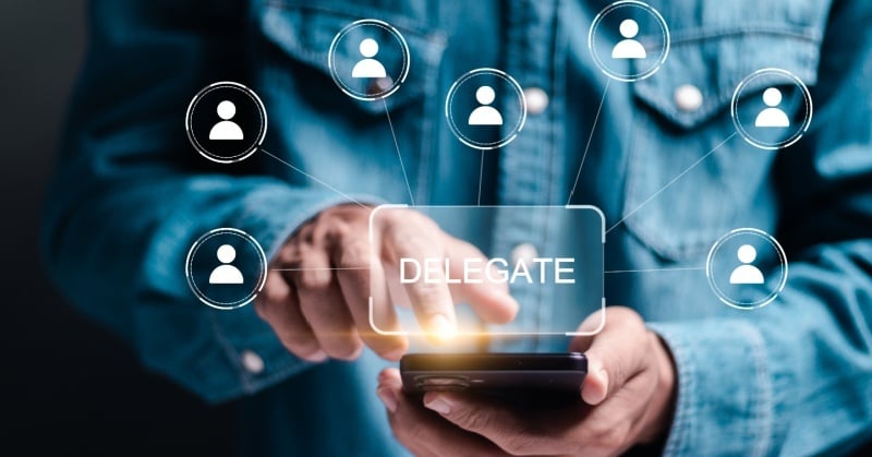 Person in a denim shirt using a smartphone, with the word “DELEGATE” displayed on screen. Around the screen are digital icons representing team members, connected by lines, symbolizing task delegation through technology.