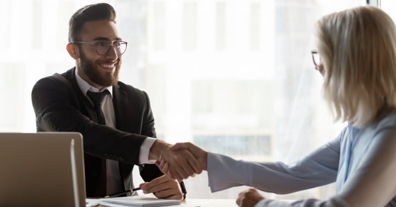 A smiling accounting job candidate in a suit shakes hands with an interviewer across a desk during a professional job interview in a bright office.