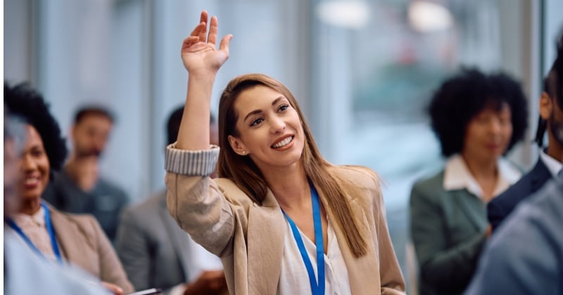 Smiling woman with long hair wearing a beige blazer and blue lanyard raises her hand in a professional conference setting, surrounded by other attendees.