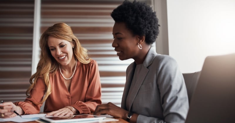 Two professional women in a business meeting reviewing financial documents at a desk, collaborating and smiling in a modern office setting.