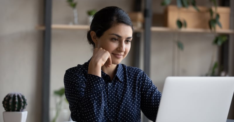 Smiling woman with dark hair wearing a navy blue, polka-dot blouse, seated at a desk and looking at a laptop screen in a modern, softly lit office with plants and wooden shelves in the background.