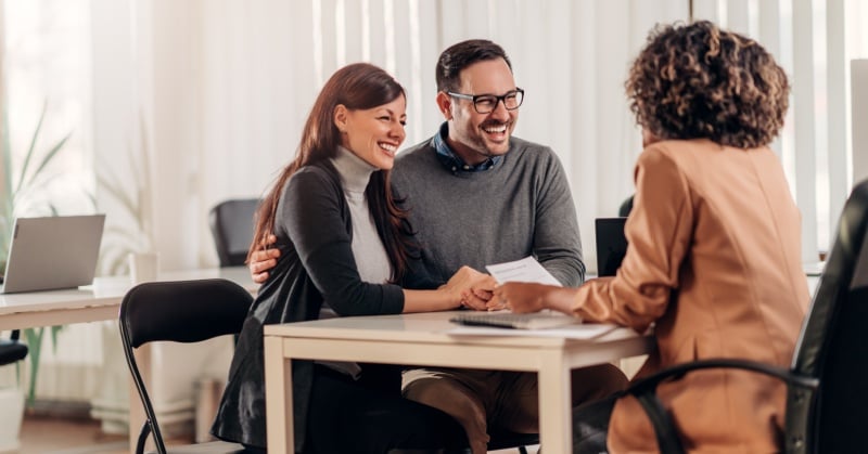 A professional financial advisor meeting with a smiling couple in a modern office setting. The advisor is handing them a document while the couple appears pleased and engaged, suggesting a positive client relationship.