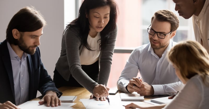 Diverse group of business professionals collaborating at a conference table with documents and notebooks during a daytime meeting in a modern office.