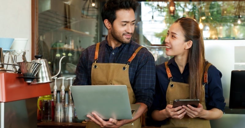Two small business owners, a man and a woman wearing aprons, stand together in a café holding a laptop and tablet, discussing their work.