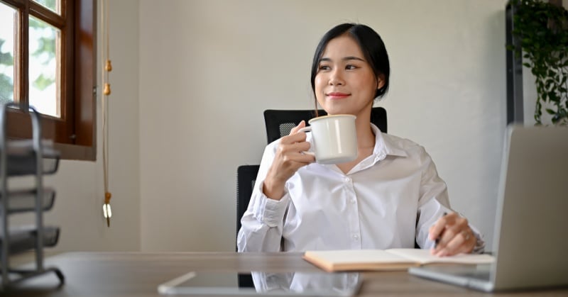 Professional woman in a white shirt smiling while holding a coffee mug at her desk, suggesting a moment of reflection during work.