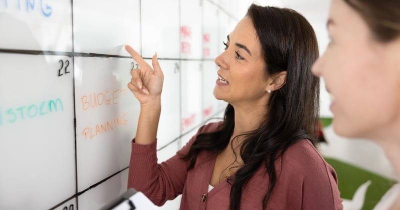 Two professionals discussing budget planning on a whiteboard calendar.