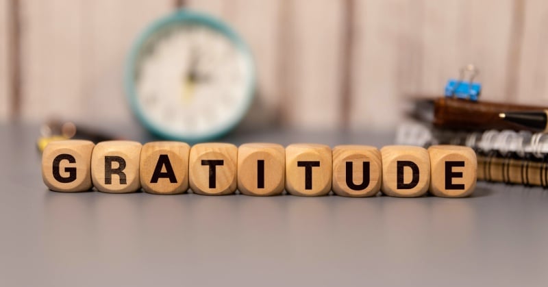 Wooden letter blocks spelling the word 'GRATITUDE' placed on a desk with a blurred clock, notebooks, and office supplies in the background.