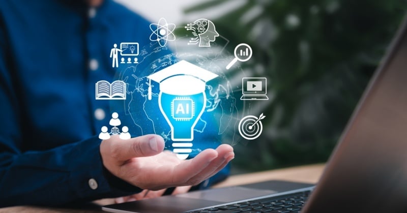 A person holding a glowing blue AI-themed lightbulb icon surrounded by digital education and technology symbols, including a graduation cap, book, video, and data chart, with a laptop in the foreground.