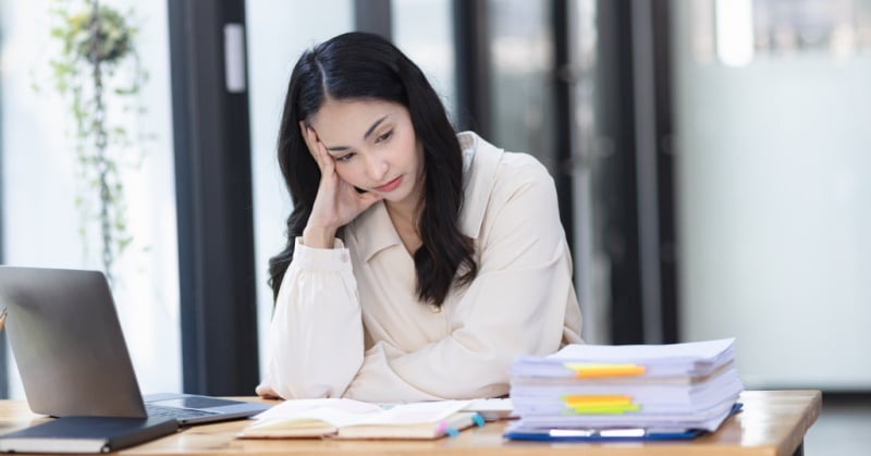 Stressed accounting professional sitting at a desk with a laptop and stacks of paperwork, appearing overwhelmed or fatigued.