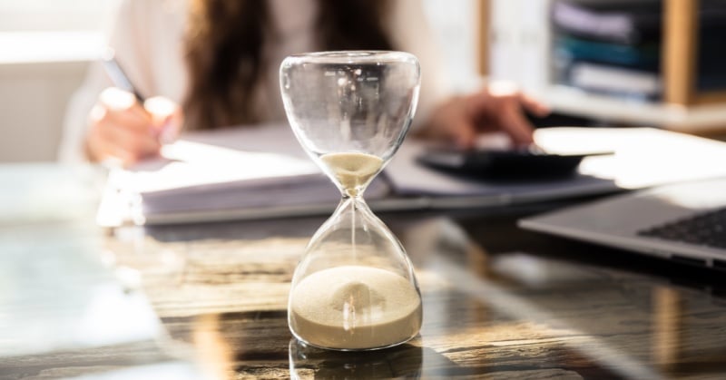 A glass hourglass with sand running through it sits on a desk in the foreground, while a blurred person works at a table with papers, a calculator, and a laptop in the background, symbolizing time pressure and year-end work.