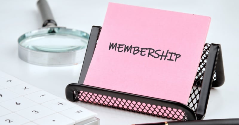 A pink notecard labeled "MEMBERSHIP" sits in a black mesh cardholder on a desk, with a magnifying glass and calculator nearby.