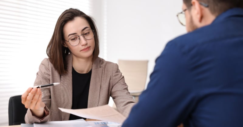 A professional accountant or advisor sits across a desk from a client, reviewing a document and gesturing with a pen while explaining details. The setting appears to be a modern office, conveying a consultative, collaborative discussion about financial or accounting matters.