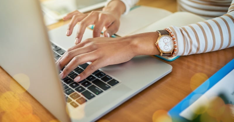 Person typing on a laptop at a desk with a notebook and documents nearby, suggesting remote work or virtual collaboration.