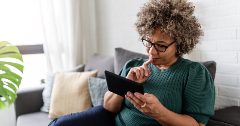 A woman with curly hair and glasses sits on a couch reading from a Kindle, appearing thoughtful and engaged.
