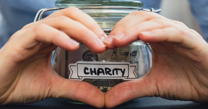 Hands forming a heart shape around a glass jar labeled "Charity" filled with U.S. dollar bills, symbolizing financial generosity.