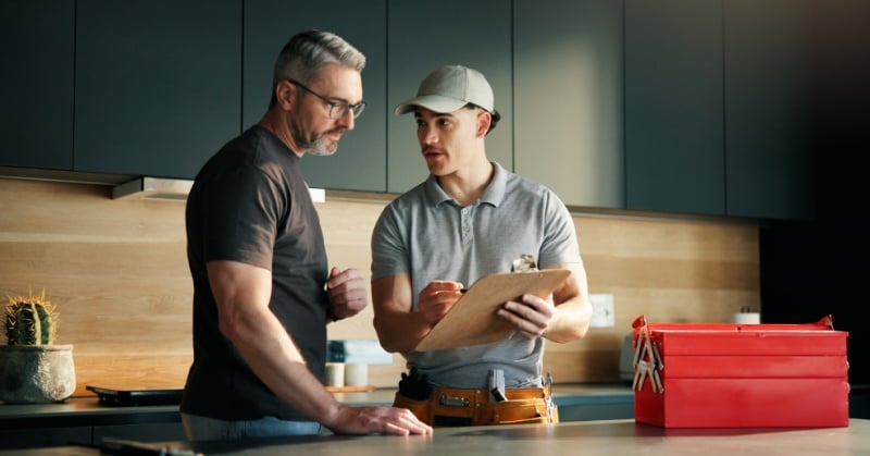Two men review job details in a modern kitchen: a trade contractor in a cap holds a clipboard while speaking with a homeowner; a red toolbox sits on the counter.