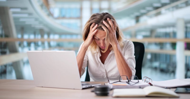 Woman at a desk holding her head in frustration while working on a laptop, with papers and glasses nearby.
