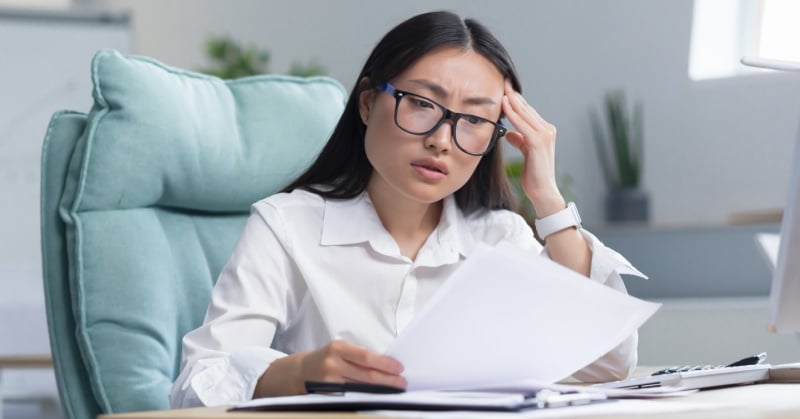 Concerned woman in glasses reviewing paperwork at her desk.