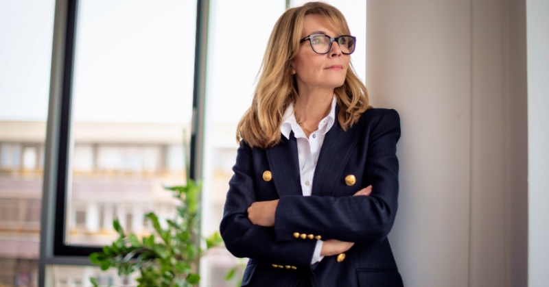 A professional-looking woman in a dark tailored blazer with gold buttons and a white blouse stands with her arms crossed, gazing to the side near a large window in a modern office setting, conveying confidence and leadership.
