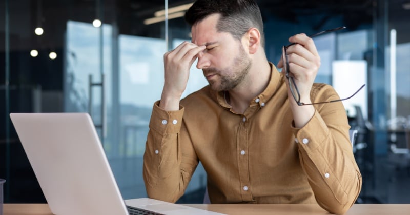Stressed office worker at a laptop, eyes closed, pinching the bridge of his nose while holding his glasses.