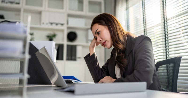 Professional woman at desk holding her head while working on a laptop in a bright office, conveying stress or fatigue.