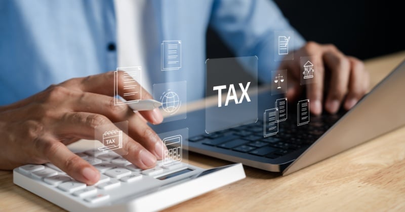 Hands typing on a laptop beside a calculator on a desk with a floating digital overlay showing TAX and document and finance icons illustrating tax season work and accounting tasks.