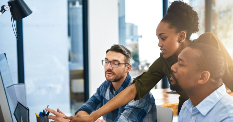 Three colleagues collaborate at a computer in a modern office, reviewing information on a large monitor.