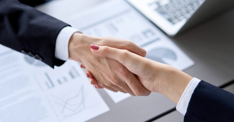 Two business professionals shaking hands over a desk with financial charts and a laptop in the background, symbolizing a business agreement or acquisition