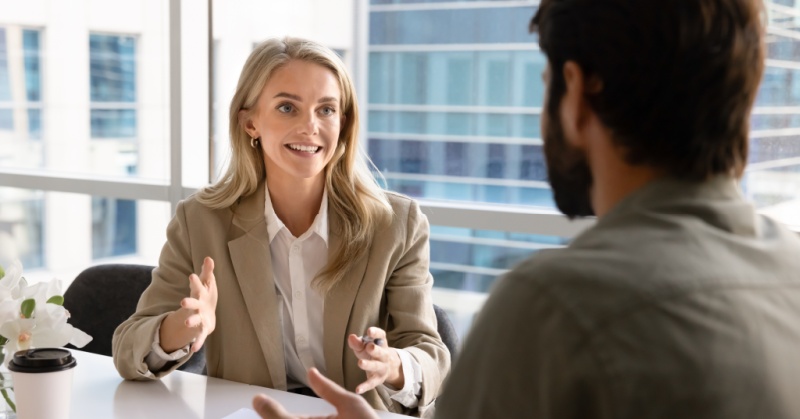 Smiling businesswoman in a beige blazer speaking and gesturing across a desk during an office meeting with a colleague.
